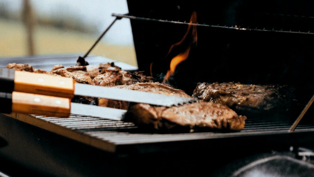 burgers being flipped using tongs on a black bbq with flames